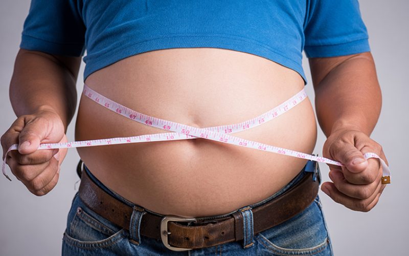 Overweight or fat adult man in very tight jeans with measuring tape on a gray background. Healthcare, medicine concept.